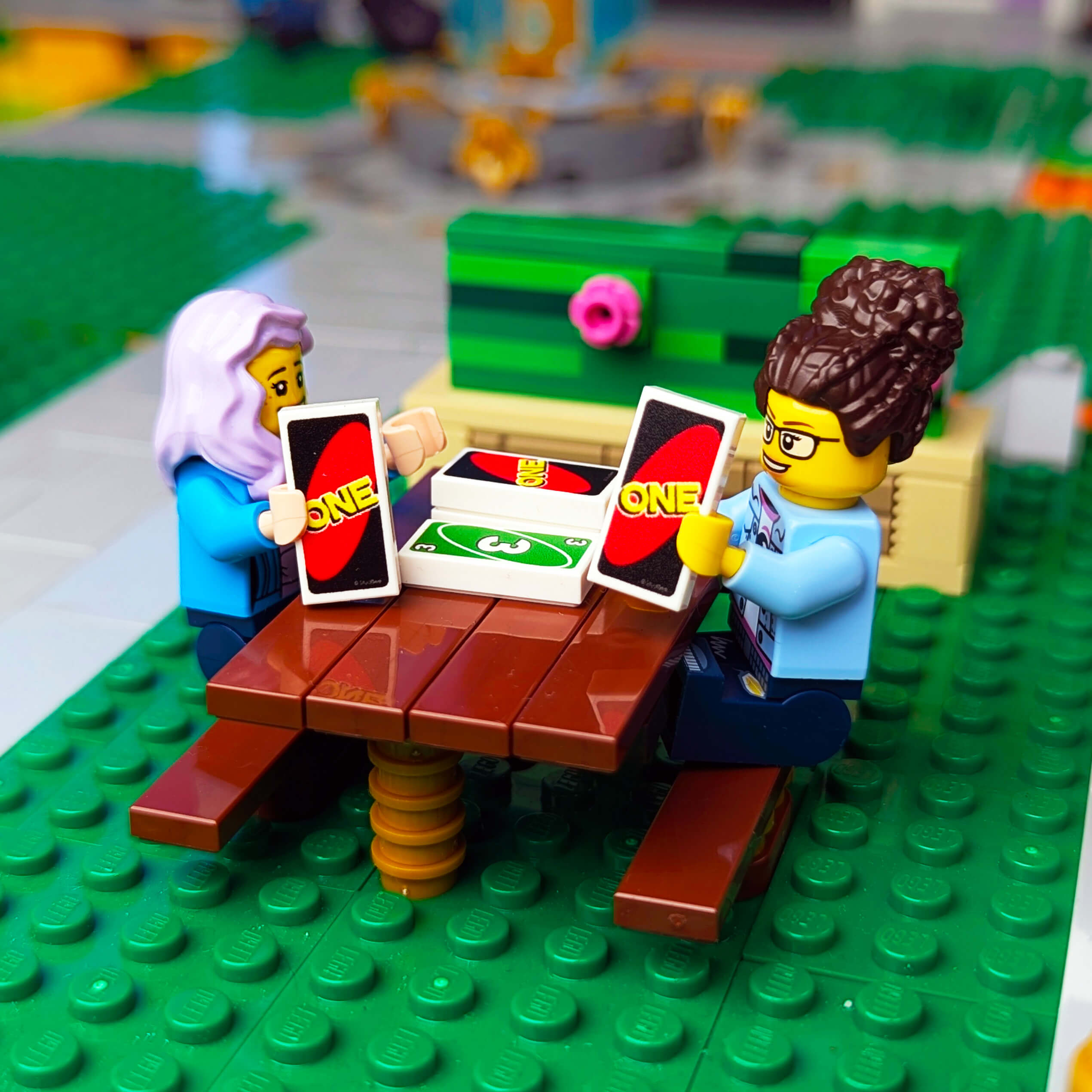 Two LEGO figures playing cards at a table on a green LEGO baseplate.