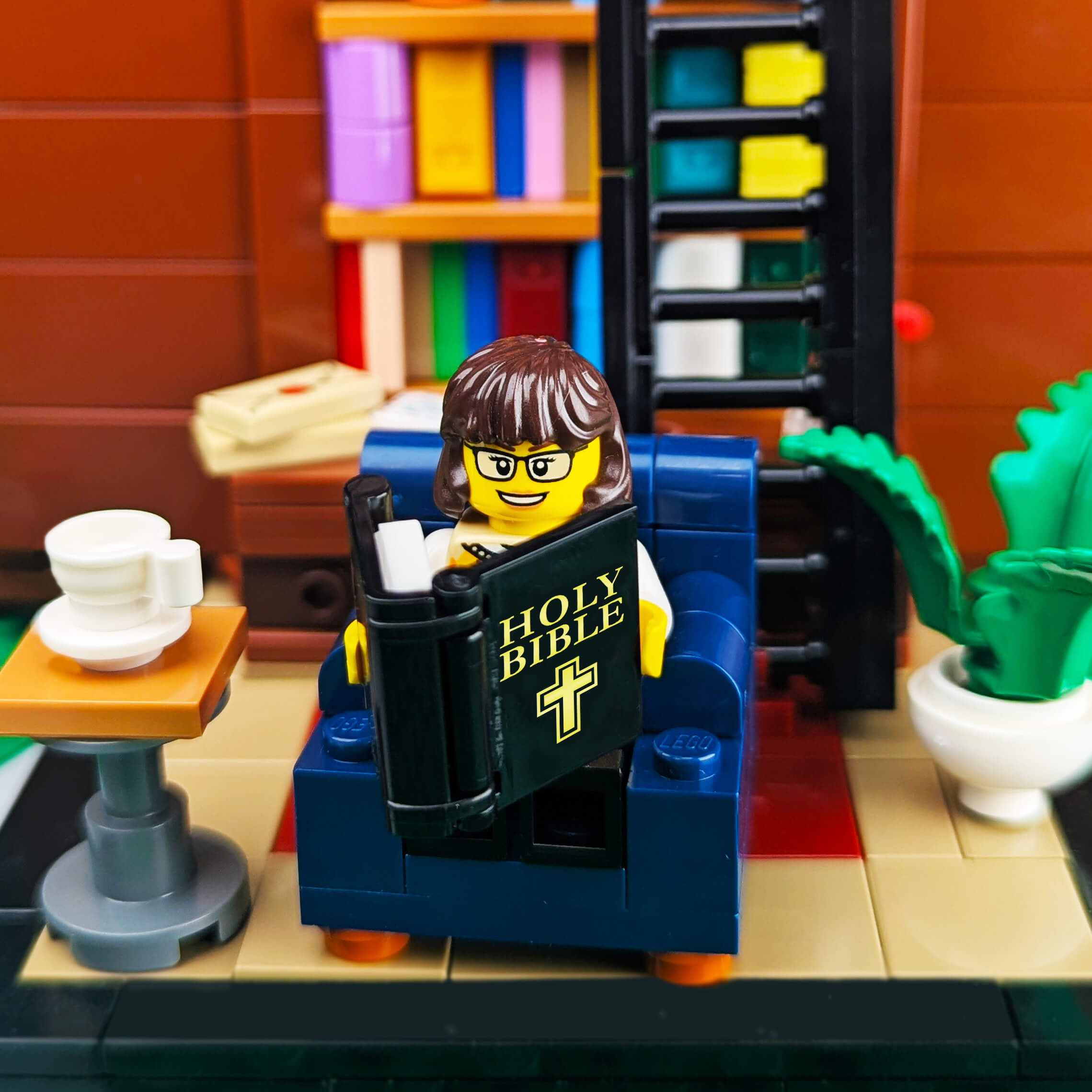 LEGO figure reading a Bible in a library setting with books and a plant.