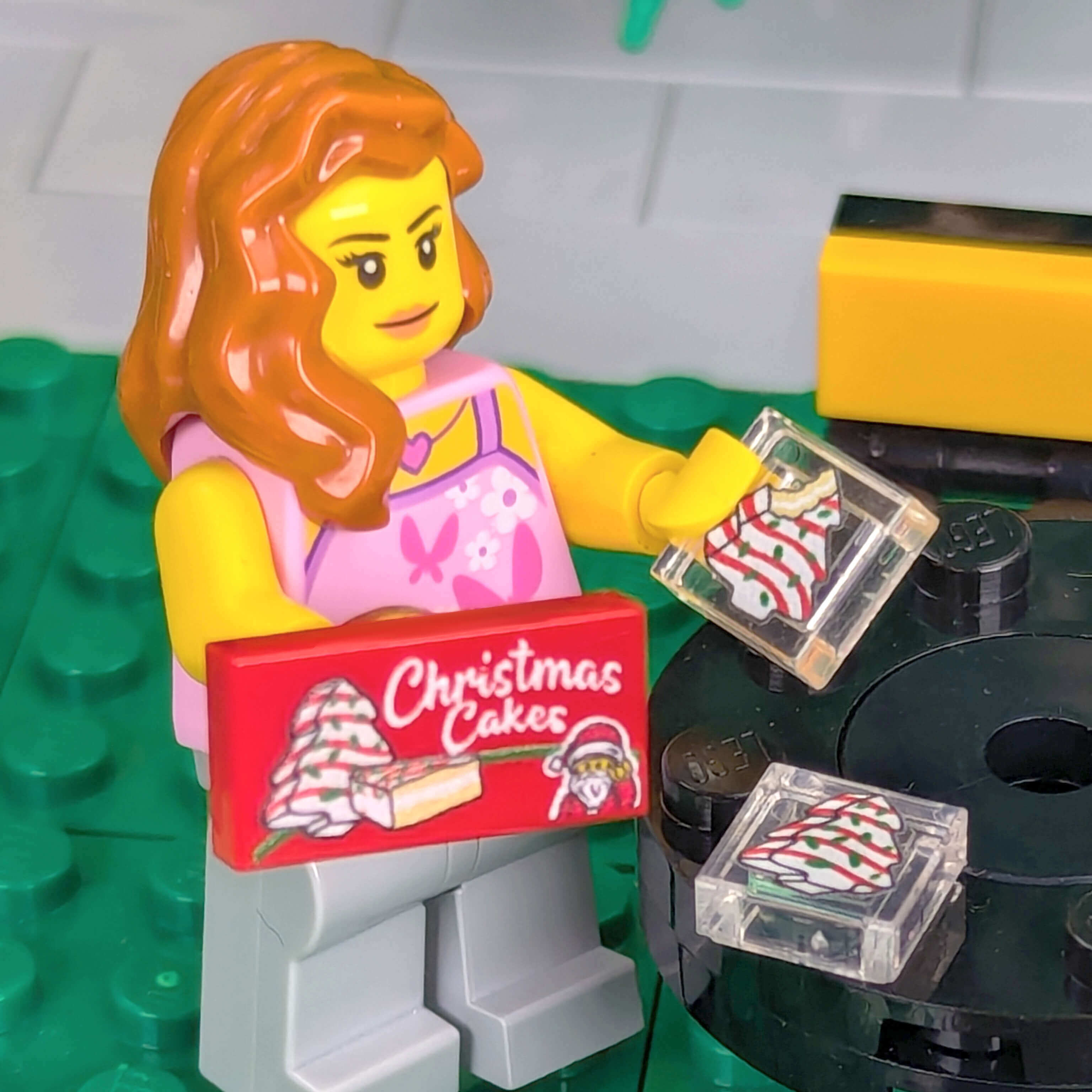 LEGO figure holding a 'Christmas Cakes' box on a green baseplate