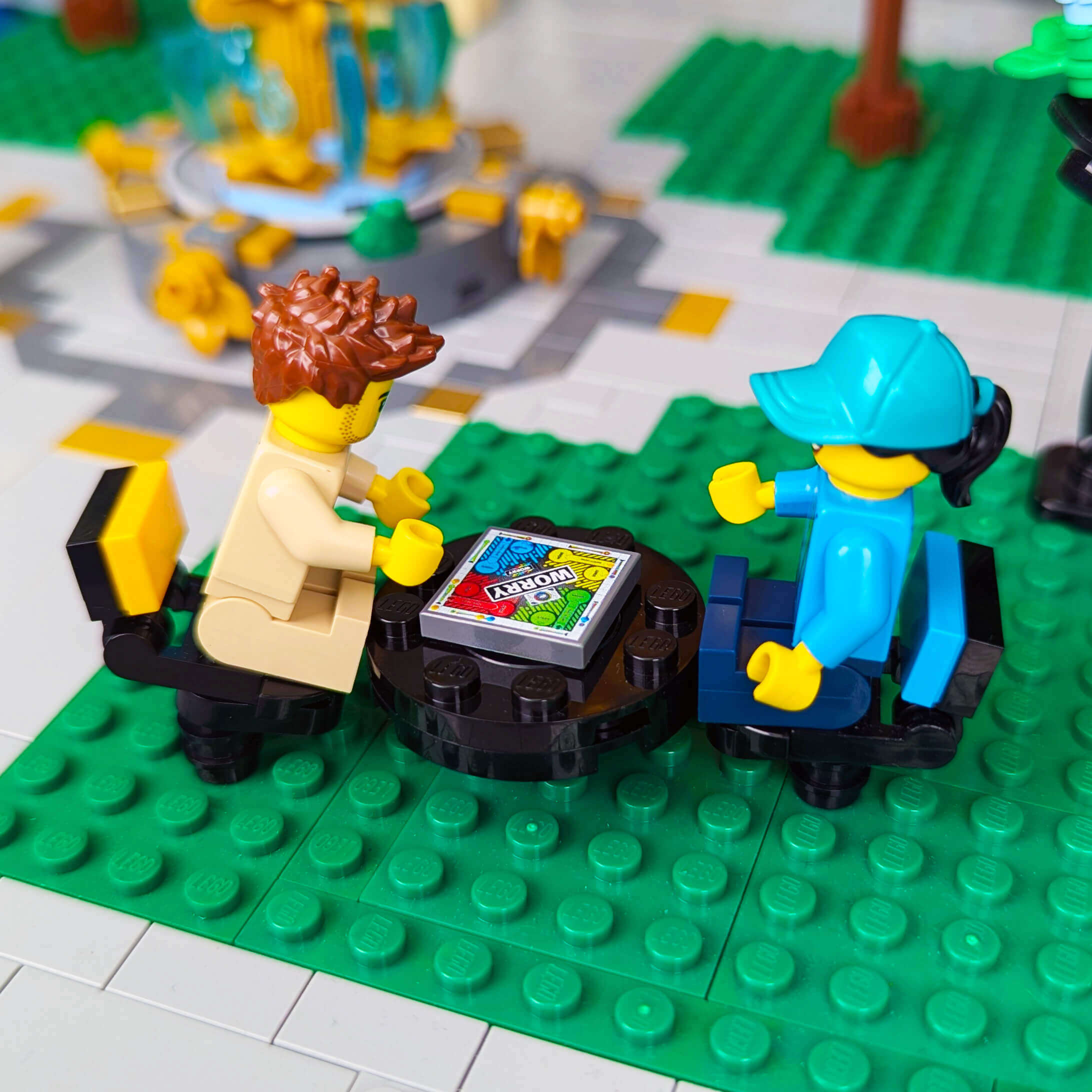 Two LEGO figures on a green baseplate playing the worry board game sitting at a table,.