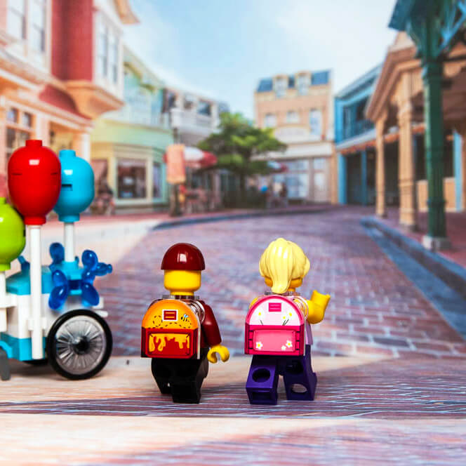 A custom printed LEGO minifig backpack featuring the character Pooh Bear Lugfly and piglet lugfly in red and yellow colors, displayed on a brick roadway with a background of simulated shops and a bicycle.