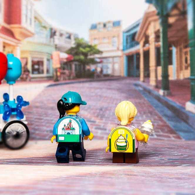 Two custom printed LEGO minifigures with a castle-themed backpack and a blue lugfly on a brick road, resembling a theme park setting.